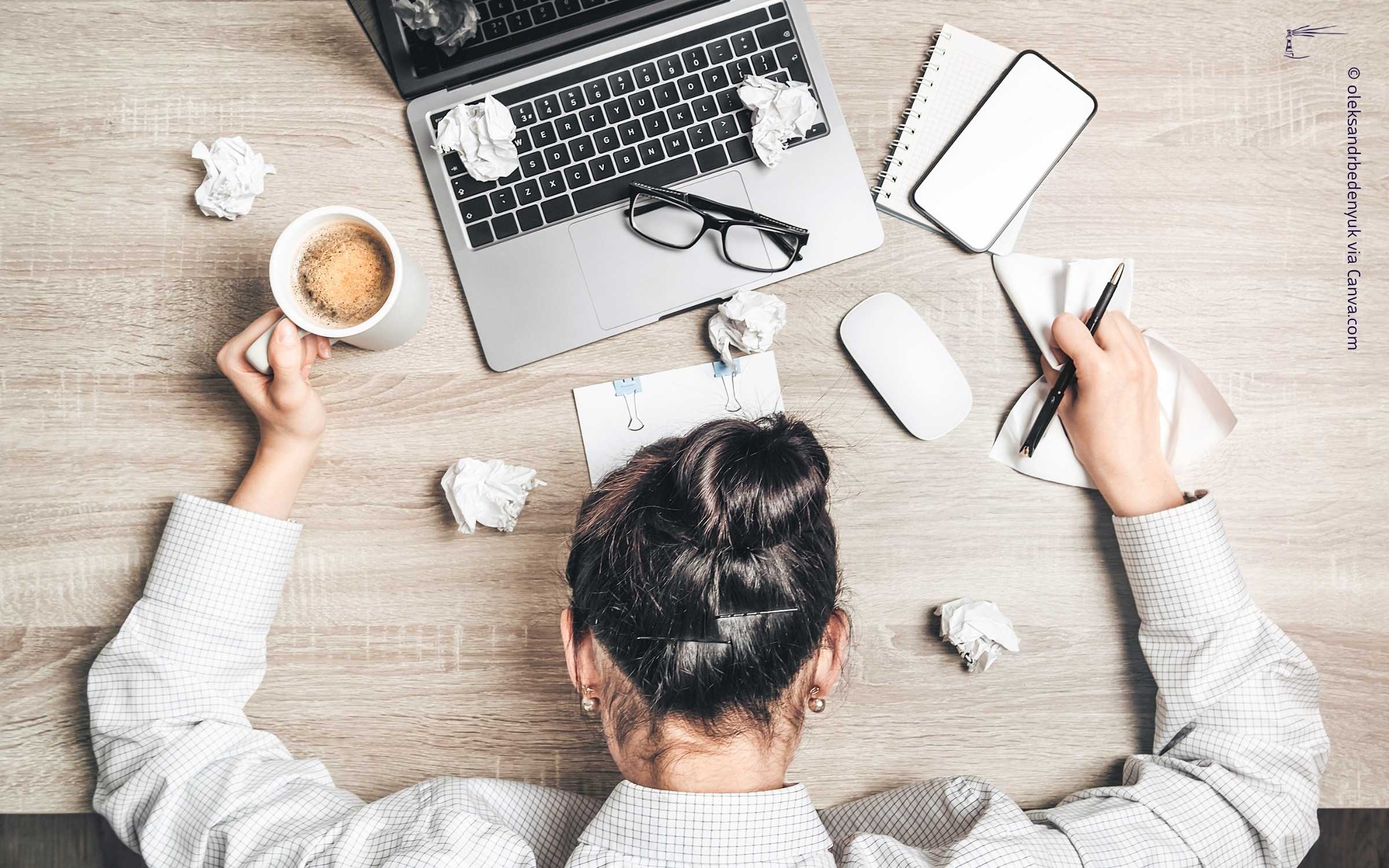 Eine Frau mit dunklen Haaren sitzt an einem Schreibtisch voller Gegenstände. Auf dem Tisch befinden sich ein silberner Laptop, ein offenes Notizbuch, zerknüllte Papiere, eine weiße Kaffeetasse, eine Brille mit schwarzem Gestell, eine Computermaus und ein blauer Kugelschreiber. Die Person hält in der linken Hand die Kaffeetasse. Es eine Atmosphäre der Überforderung als Symbol für den Beitragsinhalt zwischen Zwischen Erschöpfung und Klarheit.