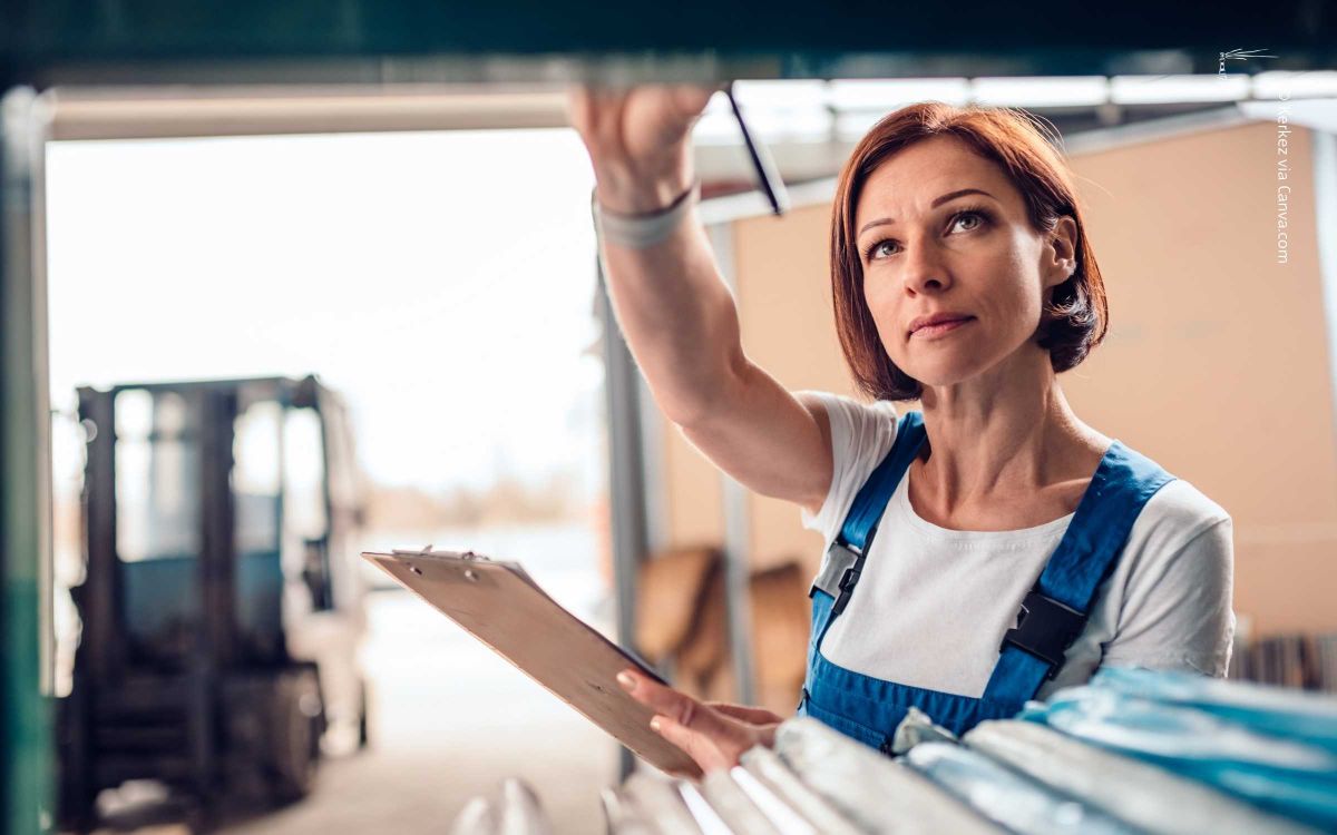 Frau mit blauer Latzhose und weißem Shirt hält Klemmbrett, greift im Lager nach Regal; unscharfer Gabelstapler im Hintergrund. Symbol für Inventur.