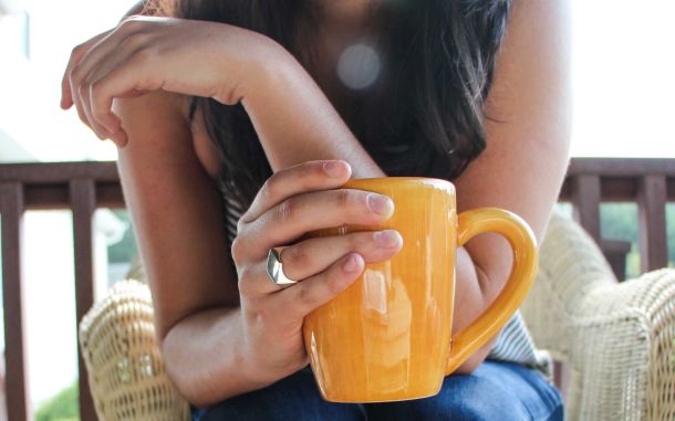 Eine Frau h&auml;lt gro&szlig;e orangefarbene Tasse in der Hand und sitzt auf einem Stuhl. Das Bild steht symbolisch f&uuml;r die Macht der Morgenroutine, die einen entspannten Start in den Tag erm&ouml;glicht.