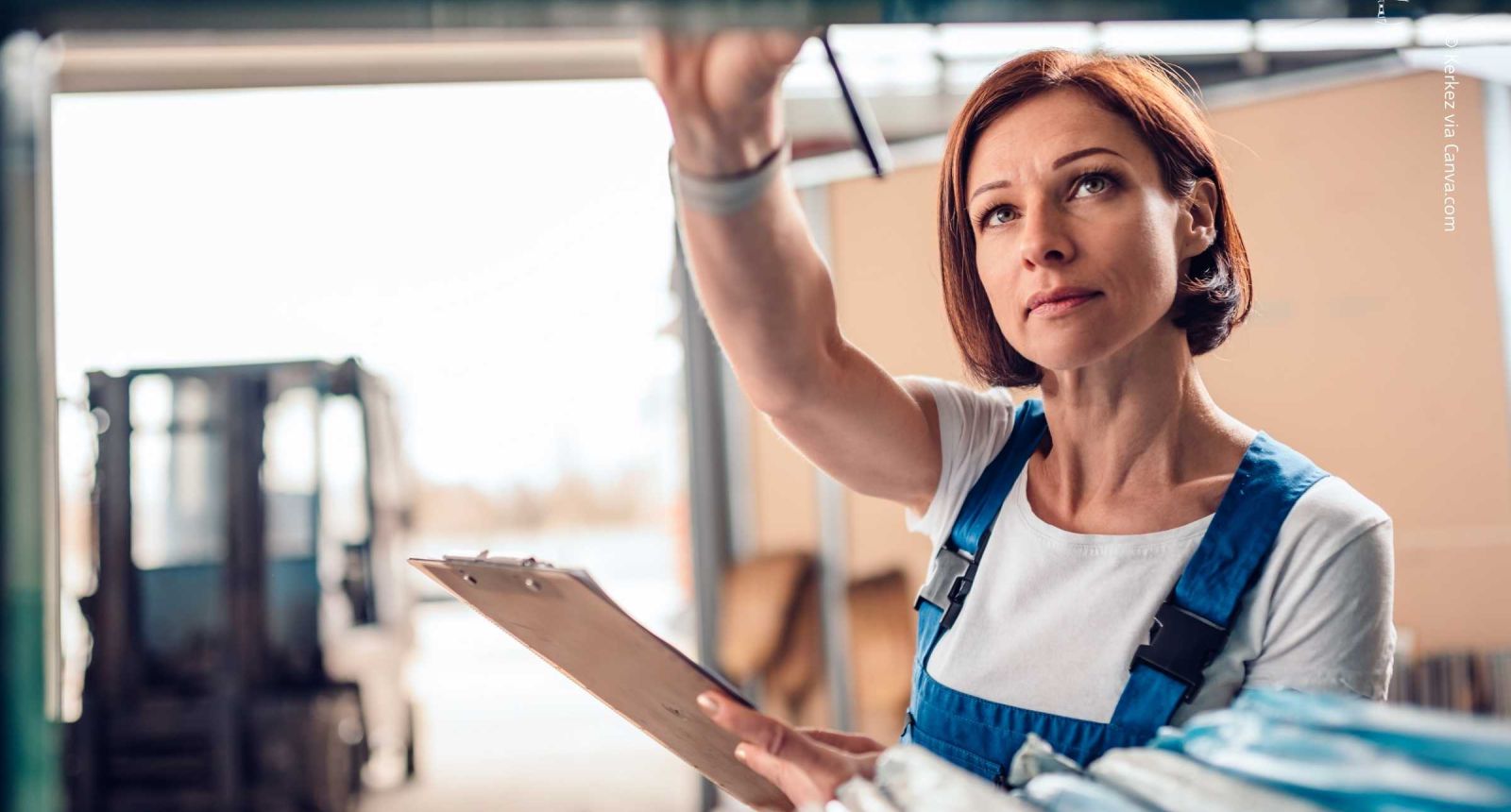 Frau mit blauer Latzhose und weißem Shirt hält Klemmbrett, greift im Lager nach Regal; unscharfer Gabelstapler im Hintergrund. Symbol für Inventur.