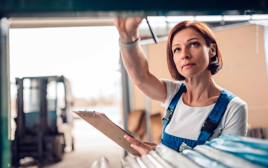 Frau mit blauer Latzhose und weißem Shirt hält Klemmbrett, greift im Lager nach Regal; unscharfer Gabelstapler im Hintergrund. Symbol für Inventur.